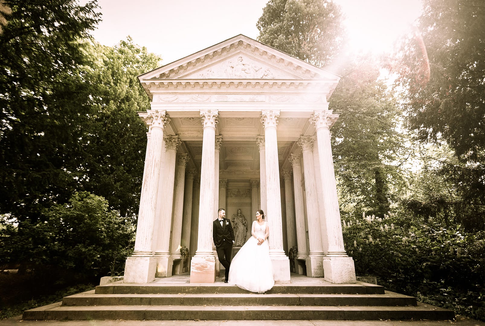 Ein Brautpaar lehnt sich an die Säulen des Minervatempels im Schwetzinger Schlossgartens an. Das Traupaar schaut sich in die Augen. Das Tempel wird rückseitig von Sonnenstrahlen gestreift.