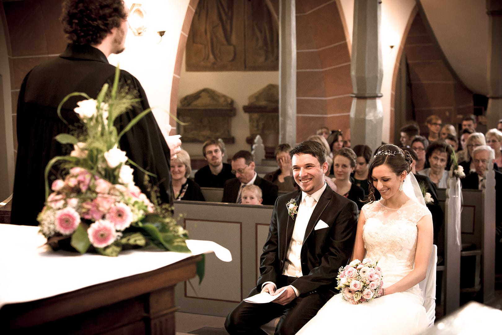 Der Priester steht vor dem Hochzeitspaar und erzählt. Die Braut und der Bräutigam lachen.