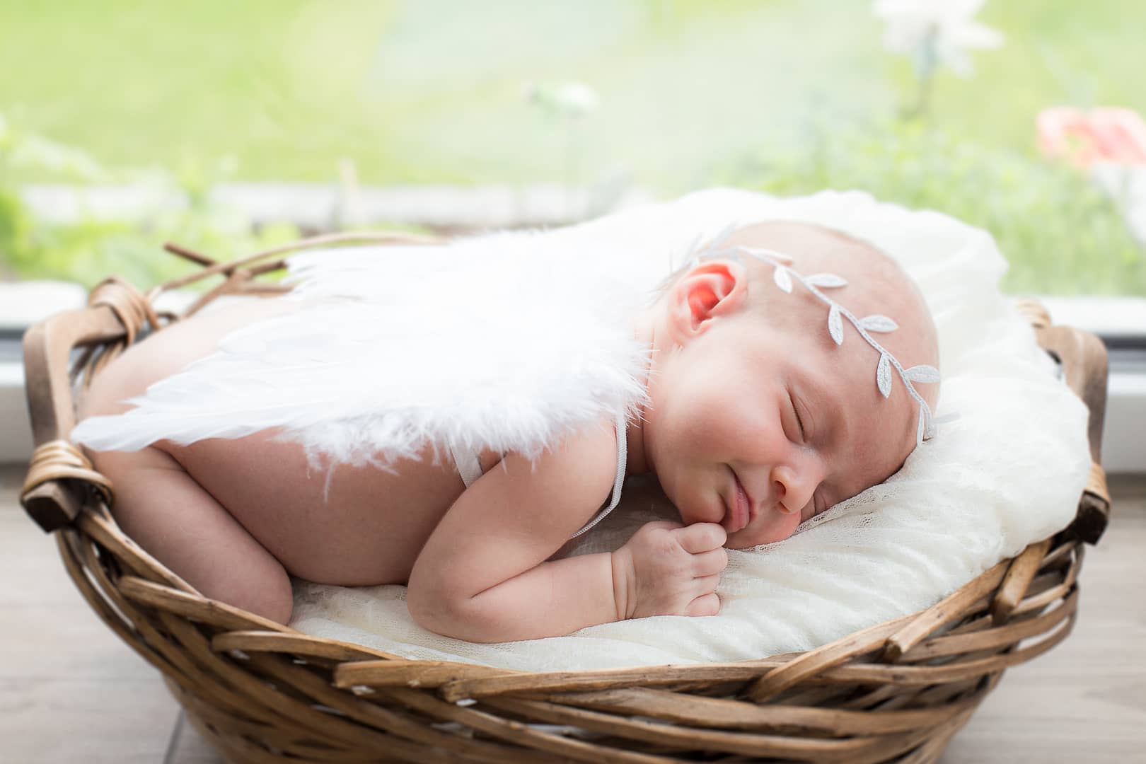 Baby mit Engelsflügeln und Babyhaarkranz schläft auf dem Bauch im kuschligem Babykorb