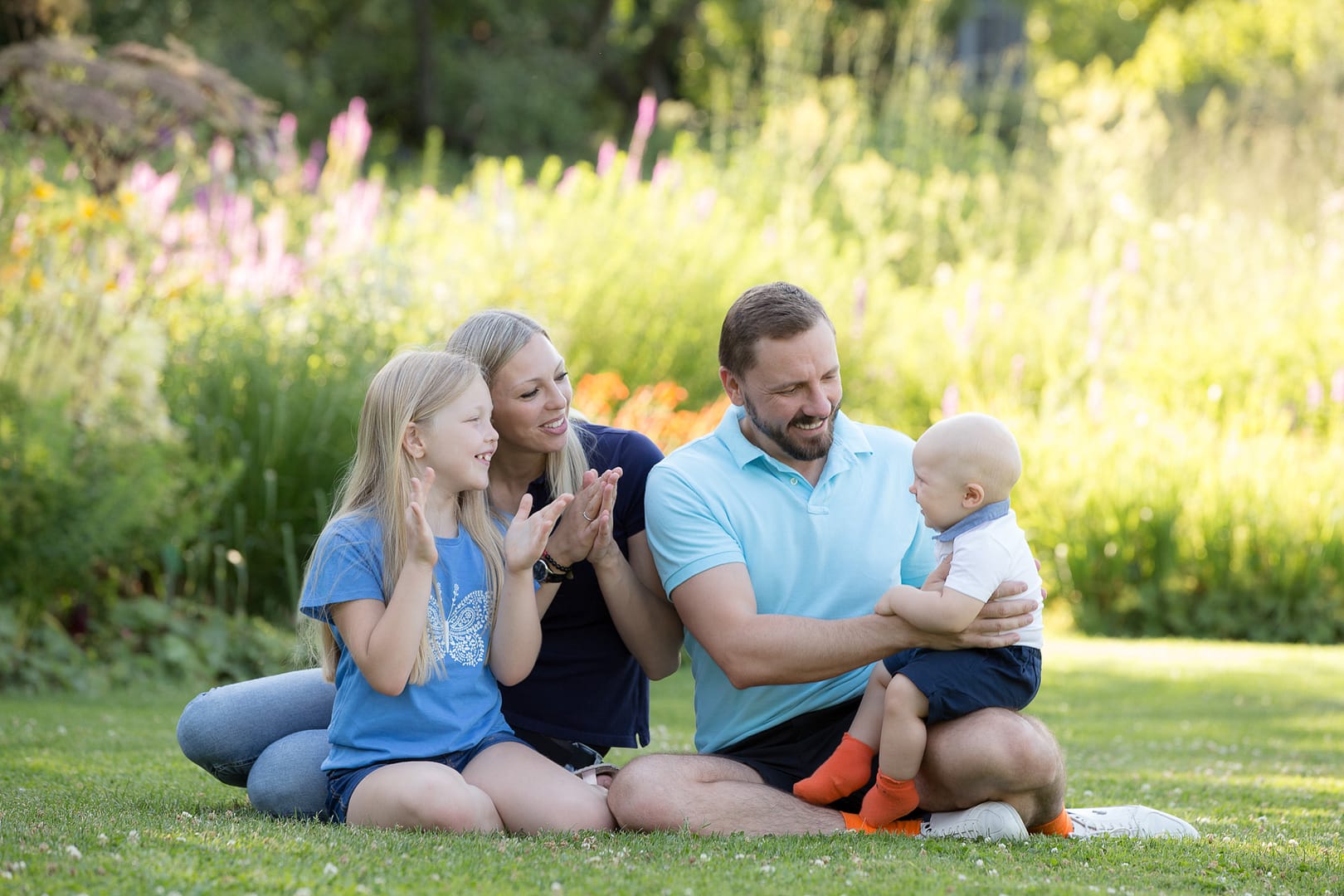 Fröhliche Familie mit Baby sitzend auf der Wiese des Weinheimer Schau- und Sichtungsgartens Hermannshof. Mama und Tochter klatschen dem Kleinkind zu. Papa hält das Baby fest.