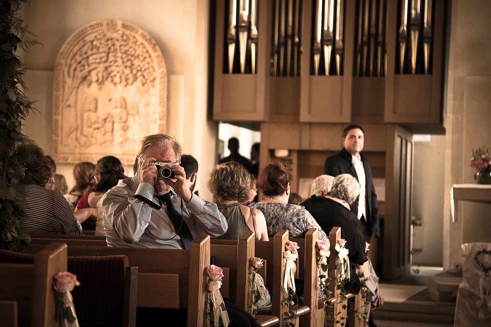 Ein Gast hat sich mit seiner Kamera in Stellung gebracht, um die Braut beim Eintritt in den Kirchensaal zu fotografieren, während im Hintergrund der Bräutigam vor dem Altar warten.