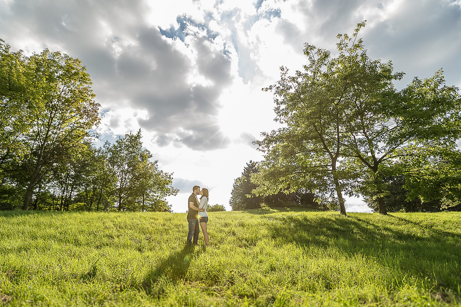 partner-fotoshooting: Ein junges Paar steht in einem hügligen Feld und küsst sich. Beidseitig sind Bäume, im Himmel sehr bewölkt und die Sonne scheint durch die Wolken hindurch.