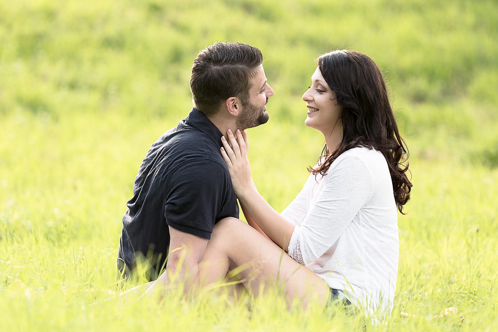partner-fotoshooting: Ein Liebespaar sitzt in einem hellgrünen Feld nah bei einander. Sie lächeln und schauen sich in die Augen. Die Frau fasst den Mann an seinem Hals an.