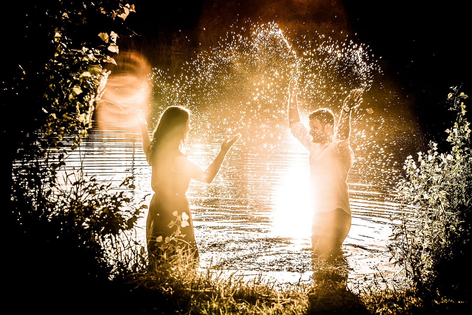 partner-fotoshooting: Ein Liebespaar steht in einem See knietief mit Abstand zu einender und schauen sich an. Sie werfen beide gleichzeitig das Wasser aus dem See hoch. Durch die Strahlen der untergehenden Sonne leuchten die Wassertropfen regelrecht.