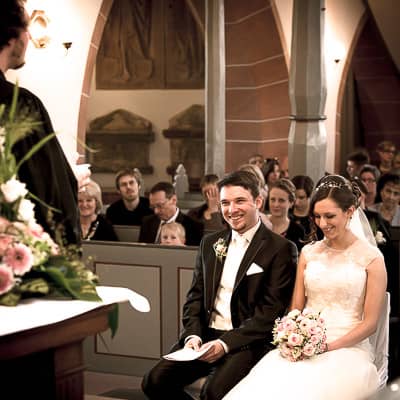 Der Priester steht vor dem Hochzeitspaar und erzählt. Die Braut und der Bräutigam lachen.