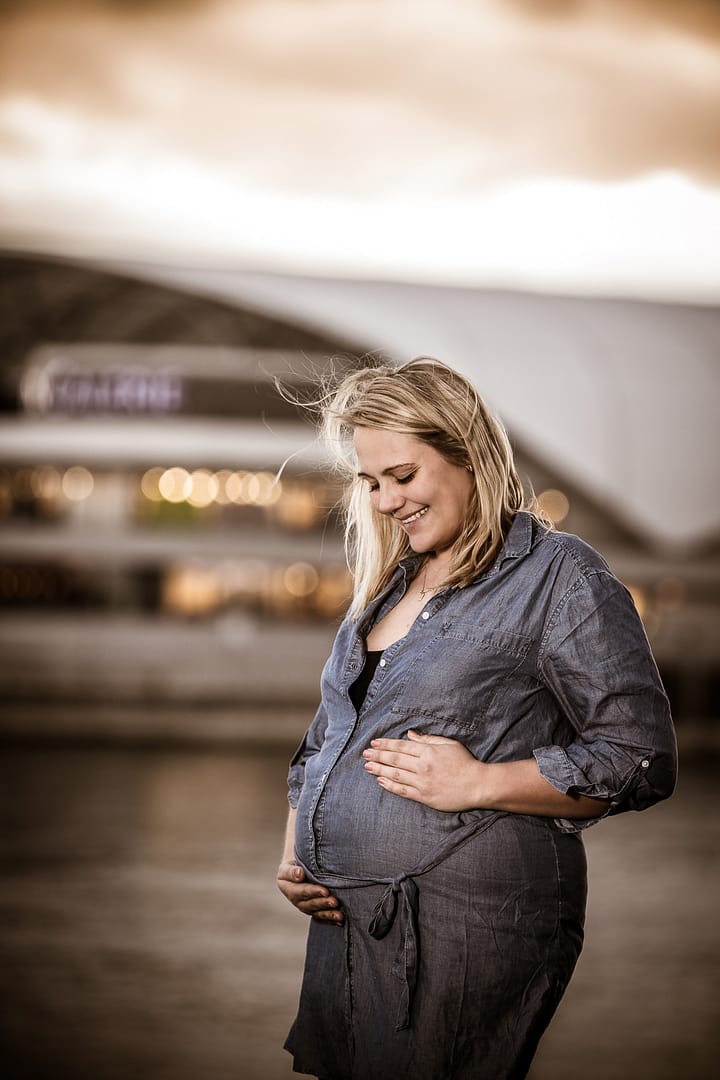 Babybauch-Fotoshooting: Eine Schwangere hält mit ihren Händen ihren Babybauch von unten und oben, während Sie zu ihrem Bauch herunterschaut und lächelt.