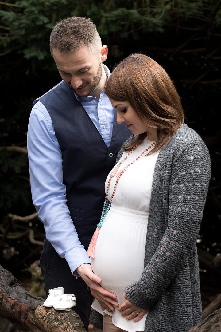 Babybauch-Fotoshooting: Eine schwangere Frau und ihr Mann stehen dicht nebeneinander und fassen beide an ihren Bauch, während beide nach unten auf zwei weiße Babyschuhe schauen, die auf einem Ast platziert sind. Sie befinden sich an einer Lichtung im Wald.