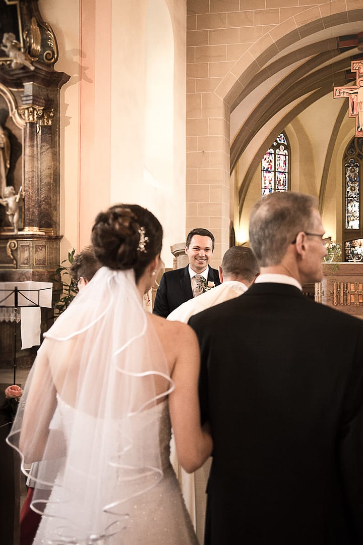 Die Braut, geführt durch ihren Vater, steht nicht mehr weit weg von dem Altar. Sie und ihr Bräutigam schauen sich gegenseitig in die Augen.