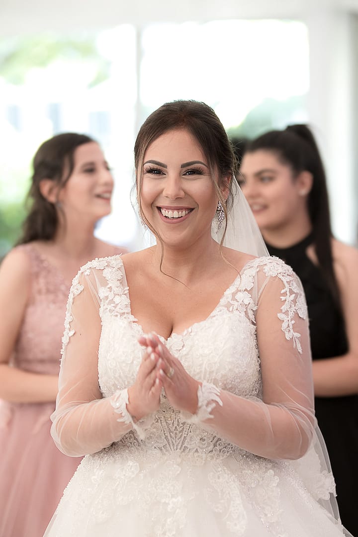 Die Braut lächelt voller Freude und hält dabei ihre Händen zusammen. Im Hintergrund sind Gäste, die ebenfalls lächeln.