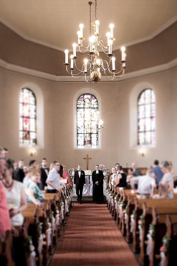 Der Bräutigam und die Priesterin stehen vor dem Altar und warten auf das Eintreten der Braut. Auch die Traugäste haben sich aufgestellt.