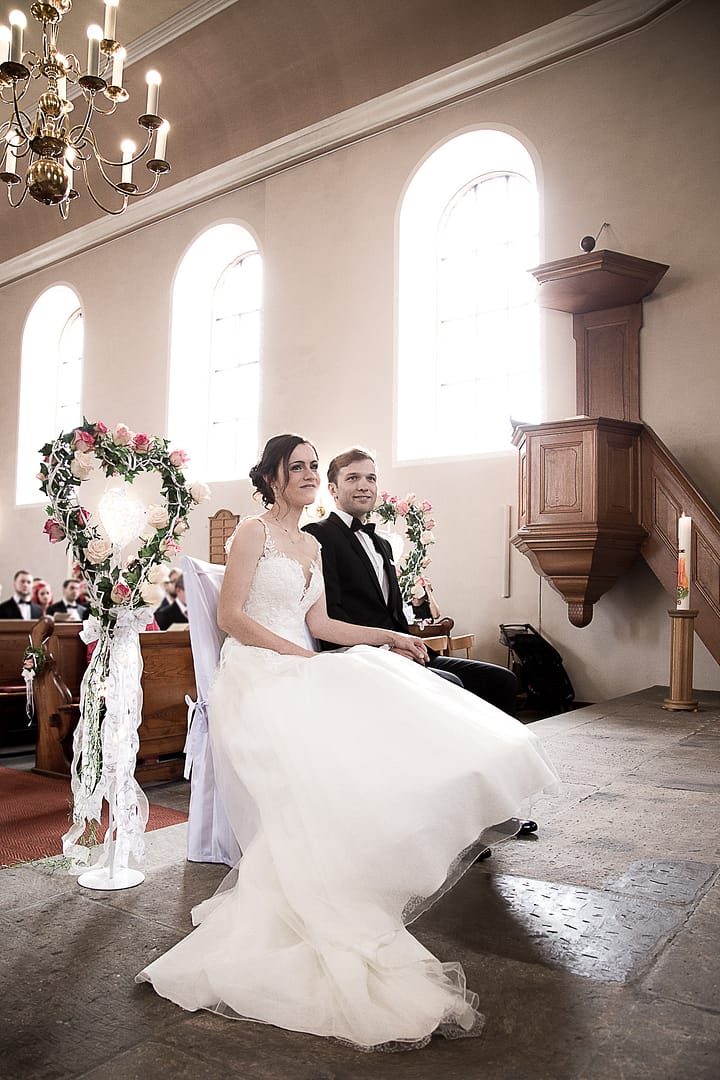 Das Hochzeitspaar sitz auf den Traustühlen nebeneinander. Sie schauen gerade aus und lächeln.