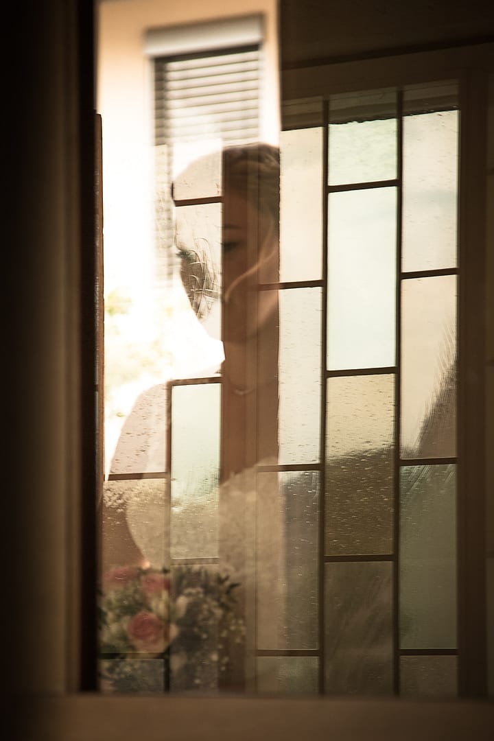 Die Braut wartet hinter der Kircheneingangstür auf ihren Eintritt in den Kirchensaal. Sie ist hinter den Gläsern der Tür teils silhouettenhaft erkennbar.