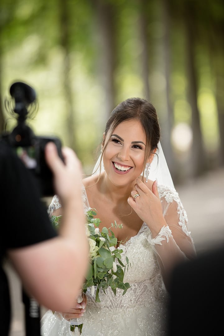 Die Braut hält ein Interview für das Hochzeitsvideo im Garten der Location ab. Sie hat ein glückliches Lächeln im Gesicht. Sie wird hierbei von einem Kameramann gefilmt.