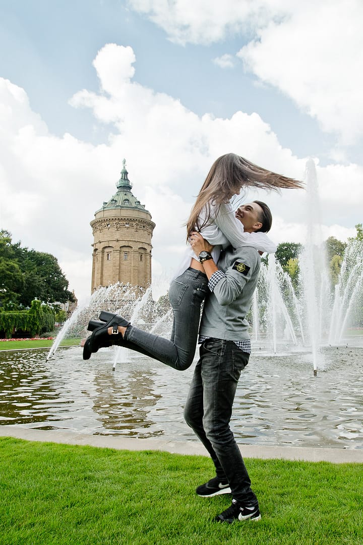 Ein junger Mann hält seine Liebste in den Armen hoch, während sie sich drehen. Sie hält sich um seinen Hals fest. Im Hintergrund sind Fontänen und der Wasserturm von Mannheim.