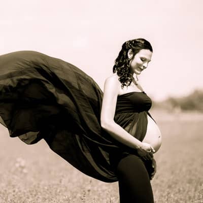 Babybauch-Fotoshooting: Schwangere Frau mit dunklem Kleid mit langem Schleier steht im Feld, streichelt mit ihrer Hand ihren nackten Babybauch und schaut dahin.